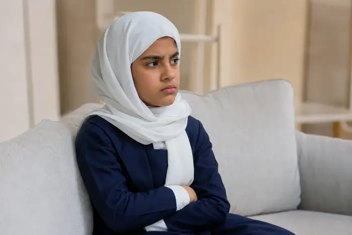 Realistic photograph, same young Khaleeji Emirati girl (same face, navy school uniform, light hijab), frustrated tense expression sitting on a couch at home, no text